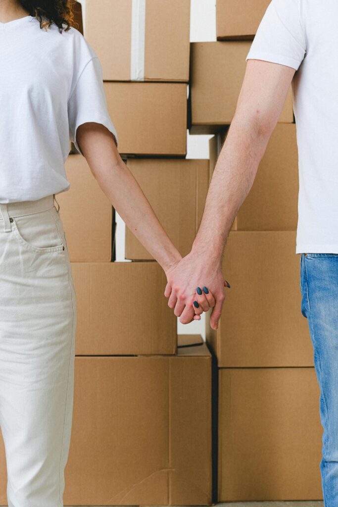 Crop unrecognizable couple in casual clothes holding hands while standing near pile of carton boxes after moving into new apartment
